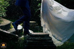 In l’Isle-sur-la-Sorgue, Vaucluse, France, under a rainy night sky, the groom gently holds the bride’s dress train as she walks down stone steps in a garden, surrounded by glistening greenery.